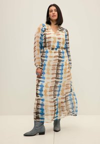 Maxi-jurk van transparant materiaal, met een tie-dye patroon in blauw, bruin en crème. Het heeft lange mouwen en een V-halslijn. Een paar grijze enkellaarsjes.