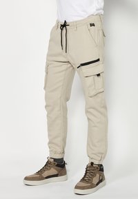 Beige Cargo-Hosen mit verstellbarem Kordelzugbund, Seitentaschen und einer Reißverschlusstasche. Kombiniert mit braunen High-Top-Sneakers.