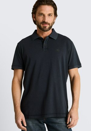 Brodaty mężczyzna o średniej długości włosach, ubrany w ciemnoniebieską koszulkę polo i niebieskie dżinsy, stojący na jednolitym jasnym tle.