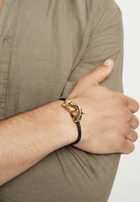 Bracelet en cuir tressé noir avec une fermeture métallique dorée au design entrelacé et un détail de surface lisse.