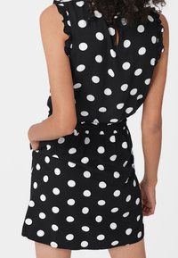 Abito nero con pois bianchi, maniche corte a volantino, vita elastica e dettaglio a chiave sul retro. Tessuto liscio con una silhouette aderente.