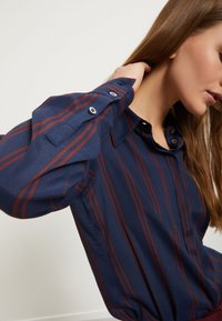 Camicia blu navy con sottili righe maroon, polsini abbottonati e colletto. Tessuto morbido e liscio con una vestibilità leggermente strutturata.