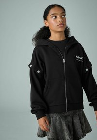 Crna dukserica na zip s kapuljačom, s srebrnim kopčama na rukavima. Nosena preko crne majice, u kombinaciji s sivom traper suknjom koja se širi.