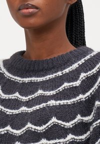 Sweter dziergany o ciemnoszarym tle, zdobiony białymi, falistymi liniami. Materiał wydaje się teksturowany i przytulny przy dekolcie.