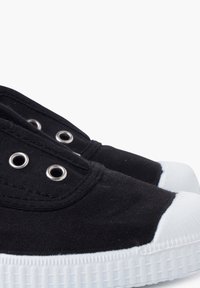 Czarne płócienne buty z metalowymi oczkami w kolorze srebrnym, białymi czubkami z gumy oraz teksturowanymi białymi podeszwami. Zbliżenie na górną część buta.