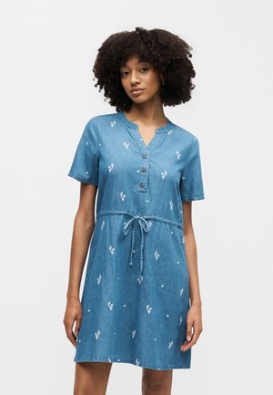 Kvinde iført en kortærmet blå denimkjole med små hvide blomsterprint og bælte i taljen, stående mod en ensfarvet hvid baggrund.