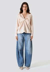 Blusa di satin rosa pallido con design anteriore attorcigliato e maniche lunghe, abbinata a jeans in denim azzurro chiaro e scarpe beige a punta.