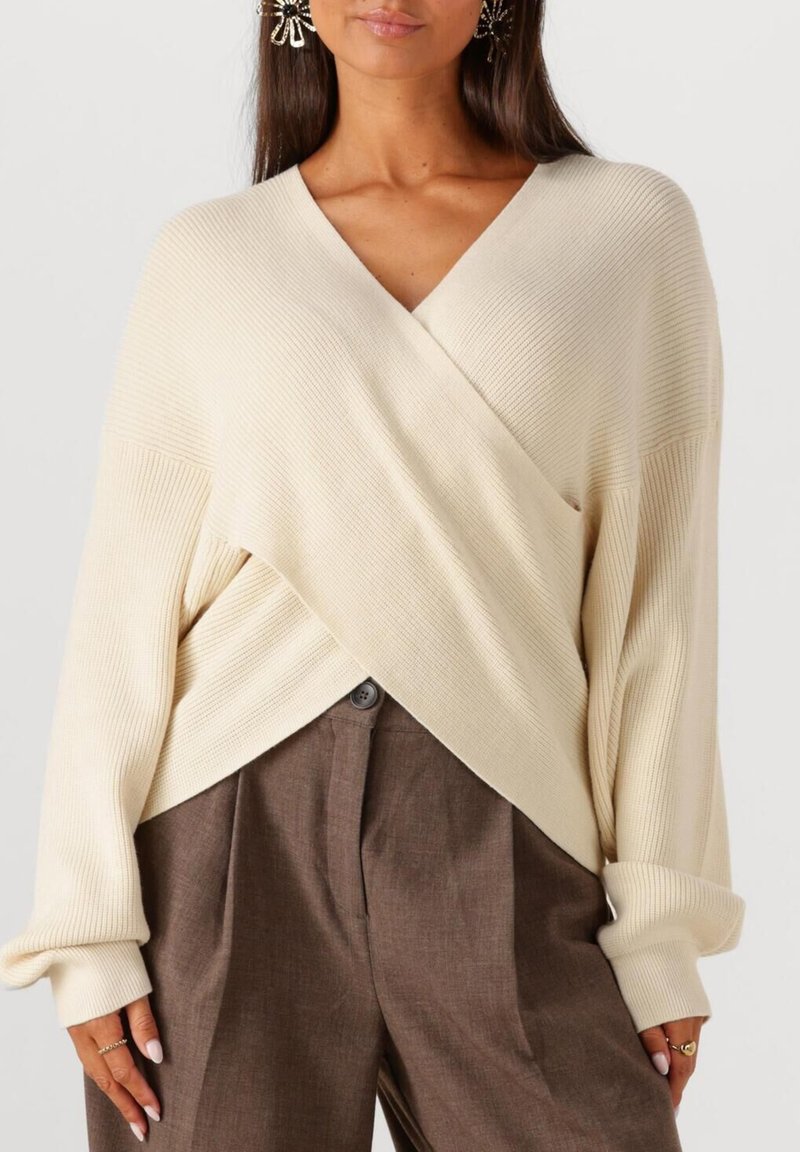 Frau in einem cremefarbenen, gerippten Wickelpullover mit langen Ärmeln und hoch taillierten, braunen, gefalteten Hosen, ergänzt durch Ringe und große Blumenohrstecker.
