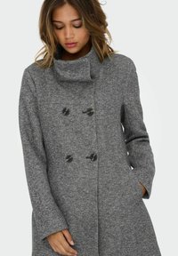Manteau gris à double boutonnage en tissu texturé, avec de grands boutons noirs et un col rabattu. Manches longues et poches latérales incluses.