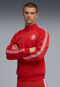 Mandlig atlet iført en rød sportsdragt med logo, med armene korslagte og et alvorligt udtryk, foran en grå baggrund. Fremhæver sport og holdsånd.