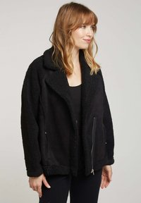 Veste en shearling noir avec un col, poche zippée à l'avant et tissu texturé. Coupe ample et manches longues.