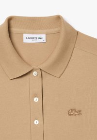 Béžová polo tričko v slim-fit strihu s textúrovanou látkou, obsahujúce trojité zapínanie na gombíky, rebrovaný golier a vyšívané logo na hrudi.