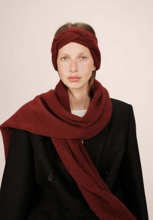 Femme à la peau claire avec des taches de rousseur, portant un bandeau en tricot rouge foncé et une écharpe assortie sur un manteau noir à double boutonnage, fond neutre.