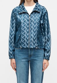 Femme portant une veste bleue zippée avec un motif géométrique en chevron et deux poches latérales à rabat, portée sur un jean bleu clair.