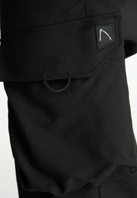 Svarta cargo-byxor med en slät yta, med en ficka med patch och cirkulär hårdvarudetalj. Designen inkluderar en broderad logotyp på fickan.