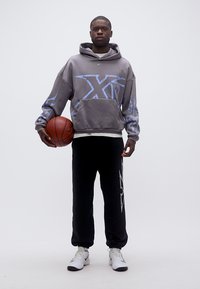 Junger Mann in grauem Hoodie und schwarzer Hose, der einen Basketball hält und vor einem schlichten weißen Hintergrund steht.