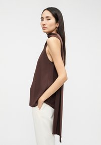 Καφέ αμάνικη μπλούζα με ντραπή σχεδίαση. Κατασκευασμένη από απαλό ύφασμα, με ασύμμετρο τελείωμα και απαλή υφή. Συνδυασμένη με ανοιχτόχρωμο παντελόνι.