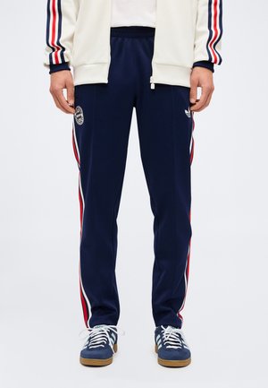 Uomo con pantaloni della tuta blu navy con strisce laterali rosse e bianche, sneakers Adidas bianche e giacca bianca con maniche a righe abbinate.