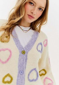 Pletený svetr v krémové barvě se vzorem srdcí v růžové, zelené a modré. Má fialový ribbovaný límec a dva knoflíky: jeden zelený a jeden modrý.