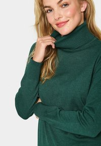 Sieviete ar gariem blondiem matiem, ģērbusies tumši zaļā augstajā kaklā, maigi turētu apkakli ar vienu roku un nedaudz smaidītu.