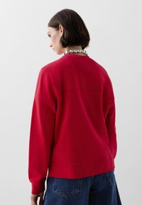 Rød sweatshirt med lange ærmer, ribbede manchetter og rund halsudskæring; har en ensfarvet tekstur og bagsøm detalje. Båret med denimshorts.