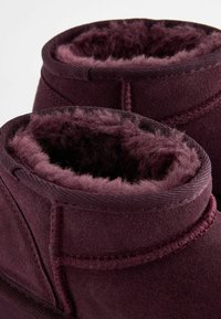 Bottes bordeaux avec une doublure douce et moelleuse. Fabriquées en daim, elles présentent un bord supérieur arrondi et des détails de couture visibles.