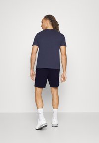 T-shirt navy à manches courtes associé à un short sombre, modèle portant des baskets blanches et des chaussettes avec un logo noir, se tenant devant un fond uni.