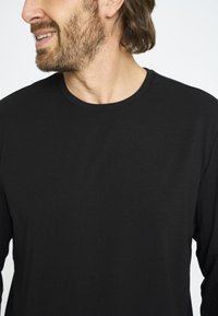 Sort langærmet skjorte lavet af ribbet stof med rund hals, der har en afslappet pasform og en glat tekstur, vist på en overkrop med lys baggrund.