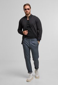 Charcoal zip-up jacket over a black t-shirt, with gray plaid trousers and white sneakers. Sunglasses complete the look, featuring sleek lines.
