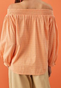 Off-the-shoulder, orange striped blouse with puffy sleeves, stretchy neckline, and textured fabric. Paired with light-colored trousers.