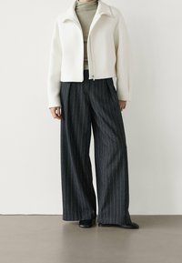 White cropped jacket with a stand collar, paired with dark pinstriped wide-leg trousers and black slip-on shoes. Smooth fabric and tailored fit.