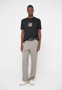 Camisa de algodão preta com um design de quadrado texturizado no peito, combinada com calças largas cinza claro e ténis brancos.