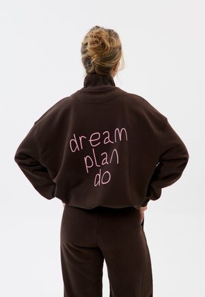 Femme avec les cheveux en chignon portant un sweat-shirt et un pantalon marron foncé, texte « dream plan do » en rose dans le dos du sweat-shirt.