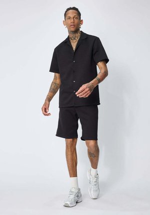 Homme avec des tatouages portant une chemise noire à manches courtes boutonnée et un short, des chaussettes blanches et des baskets blanches, marchant devant un fond blanc uni.