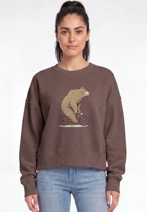 Frau mit dunklem Pferdeschwanz, die einen braunen Pullover mit einer Grafik eines Bären auf einem Fahrrad und hellblaue Jeans trägt.