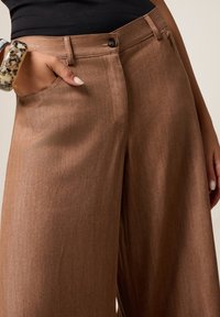 Donna che indossa pantaloni marroni a vita alta e gamba larga con bottone e cerniera, mano in tasca e bracciale tartaruga al polso.