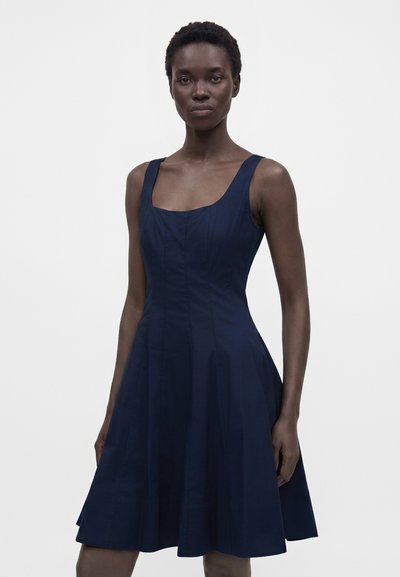 Femme aux cheveux courts portant une robe bleu foncé sans manches avec un col carré, debout devant un fond clair uni.