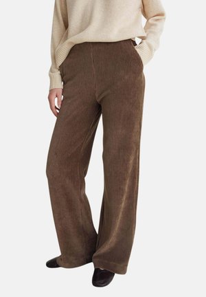 Persona que lleva pantalones marrones de pana de pierna ancha, suéter beige y zapatos negros, de pie con una mano en el bolsillo contra un fondo liso.