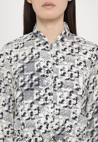 Cămașă cu guler, având un fundal alb cu un model abstract negru. Dispune de o închidere cu butoane și are o textură moale, potrivită pentru straturi.