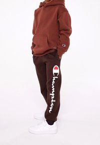 Felpa marrone con tasca frontale e logo sulla manica, abbinata a pantaloni della tuta marroni con testo e emblema "Champion" bianchi. Sneakers bianche.