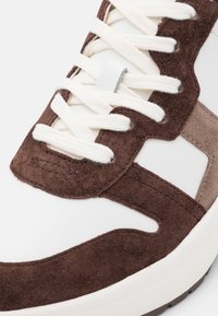 Hnědé semišové a bílé kožené tenisky s krémovými tkaničkami, které mají výrazné panely a prošívané detaily, podtržené textura designem.
