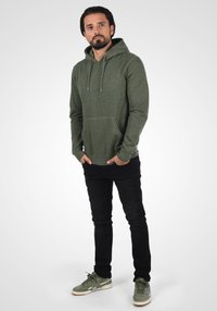 Green hoodie with a front pocket, drawstring hood, and ribbed cuffs, paired with black jeans and green sneakers.