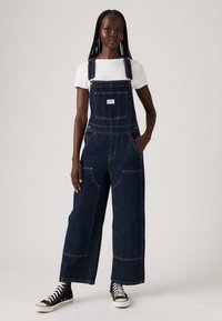 Mujer vestida con un overol de mezclilla oscura de pierna ancha, una camiseta blanca y zapatillas altas negras, de pie contra un fondo claro y liso.