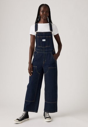 Kvinne iført mørk denim vid-legged overaller med en hvit t-skjorte og svarte høye sneakers, stående mot en enkel lys bakgrunn.