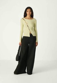 Nainen, jolla on päällään vaaleankeltainen ribattu neulepaita, jossa on napit toisella puolella, mustat leveälahkeiset housut, riipuskorvakorut ja joka pitää mustaa käsilaukkua.