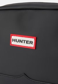 Čierna nepremokavá taška s bielou nálepkou s textom "HUNTER" orámovaným červenou farbou, detailným šitím a hladkým matným povrchom.