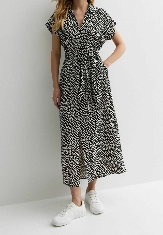 Femme portant une robe midi noire avec des motifs carrés blancs, manches courtes, une ceinture à la taille et des baskets blanches, debout devant un fond uni.
