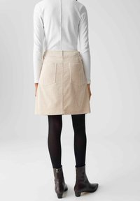 Beige corduroy rok met twee achterzakken, gecombineerd met zwarte panty en enkelboots. De top heeft een hoge halslijn en lange mouwen.