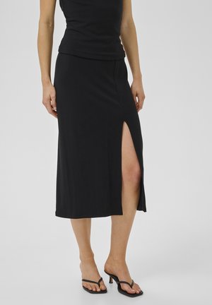 Kvinde iført en sort midi-nederdel med en højt skåret slids foran på låret, sort ærmeløs top og sorte sandaler med hæl mod en ensfarvet lys baggrund.