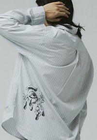 Striped long-sleeve shirt in light gray with a loose fit. Features a graphic design of an eye and drips on the side, and a classic collar.
